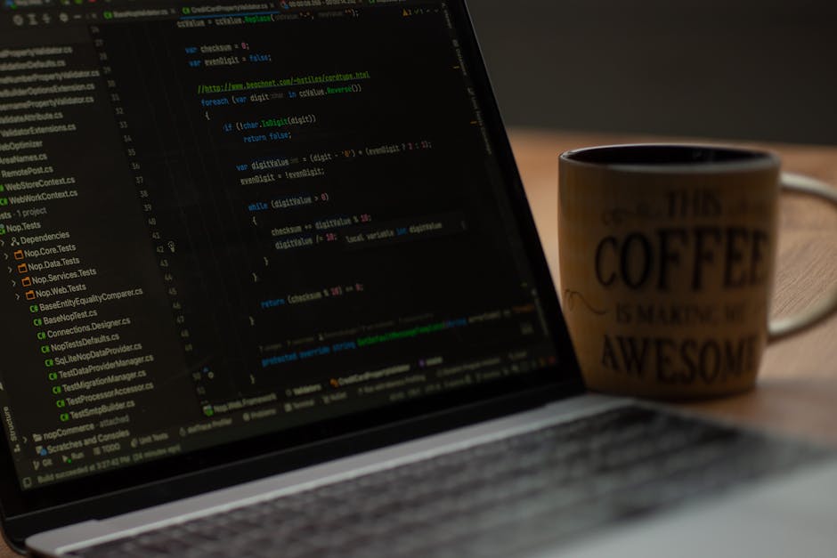 A close-up view of a laptop computer displaying lines of code on its dark screen, placed on a wooden surface. To the right of the laptop, there is a ceramic mug with a cream-colored glaze featuring bl