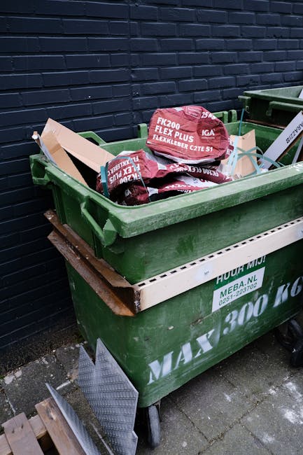 A large green wheeled skip bin positioned outdoors on a paved surface beside a dark brick wall. The bin is filled with red and white packaged garden or building materials, such as bags of fertiliser o
