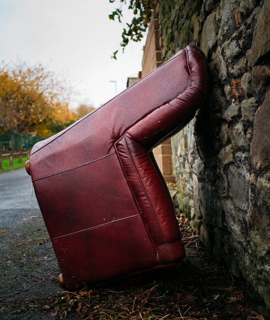 A large green wheeled skip bin positioned outdoors on a paved surface beside a dark brick wall. The bin is filled with red and white packaged garden or building materials, such as bags of fertiliser o