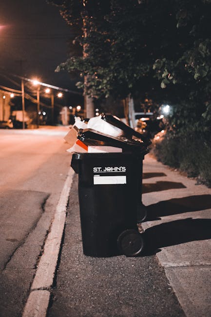 A black waste bin with a white label reading 'ST. JOHN'S' is positioned on the edge of a pavement beside a residential street at night. The bin is partially open, with various discarded items such as